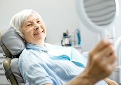 An older woman looking at her smile in the mirror while seated in the dentist’s chair after receiving her dentures in Harrisonburg