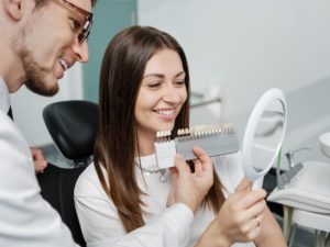 Woman with brown hair in dental chair smiling in a mirror while the dentist holds up a veneer shade guide